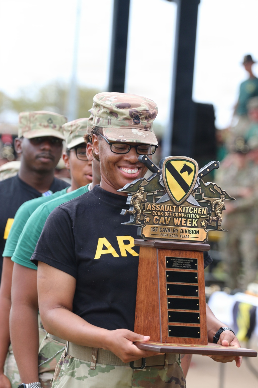 A U.S. Army soldier wearing an OCP (Operational Camouflage Pattern) uniform cap and black Army PT shirt is smiling while holding a large trophy. The trophy is for the 'Assault Kitchen Cook Off Competition - CAV Week' associated with the 1st Cavalry Division at Fort Hood, Texas. Several other soldiers in uniform are visible in the background.