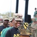 A U.S. Army soldier wearing an OCP (Operational Camouflage Pattern) uniform cap and black Army PT shirt is smiling while holding a large trophy. The trophy is for the 'Assault Kitchen Cook Off Competition - CAV Week' associated with the 1st Cavalry Division at Fort Hood, Texas. Several other soldiers in uniform are visible in the background.