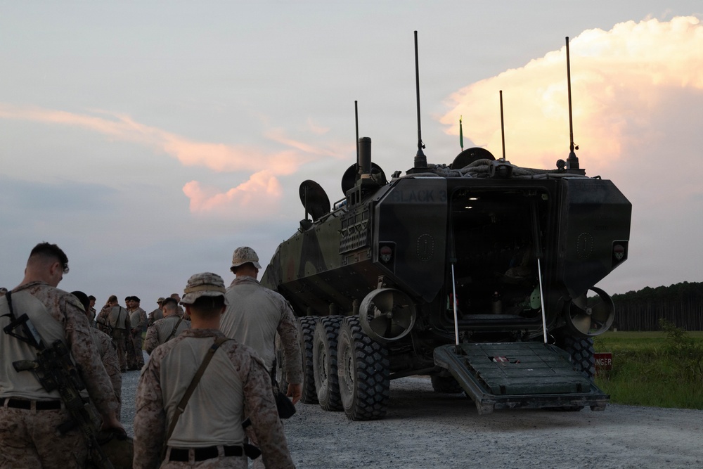 A group of military personnel in desert camouflage uniforms are gathered around a large wheeled armored vehicle, likely an AAV (Amphibious Assault Vehicle) or LAV (Light Armored Vehicle), with its rear ramp lowered. The scene appears to take place at dusk on a gravel road near a treeline, suggesting a training exercise or operational staging area. Multiple soldiers are approaching or observing the vehicle from the rear.