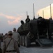 A group of military personnel in desert camouflage uniforms are gathered around a large wheeled armored vehicle, likely an AAV (Amphibious Assault Vehicle) or LAV (Light Armored Vehicle), with its rear ramp lowered. The scene appears to take place at dusk on a gravel road near a treeline, suggesting a training exercise or operational staging area. Multiple soldiers are approaching or observing the vehicle from the rear.