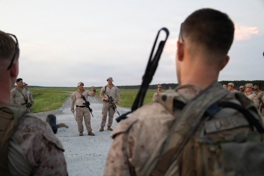 A group of military personnel in desert camouflage uniforms are gathered on a gravel road in an open field or training area. Two individuals appear to be addressing the group, with one gesturing as if giving instructions or a briefing. The setting suggests a field training exercise or pre-mission briefing at dusk.