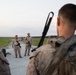 A group of military personnel in desert camouflage uniforms are gathered on a gravel road in an open field or training area. Two individuals appear to be addressing the group, with one gesturing as if giving instructions or a briefing. The setting suggests a field training exercise or pre-mission briefing at dusk.