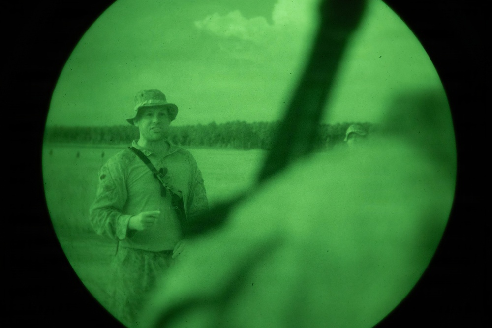 Image captured through a night vision device showing a military personnel in the field. The circular green-tinted view is characteristic of Generation 2 or 3 night vision optics. A second figure is partially visible in the background to the right.