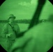 Image captured through a night vision device showing a military personnel in the field. The circular green-tinted view is characteristic of Generation 2 or 3 night vision optics. A second figure is partially visible in the background to the right.