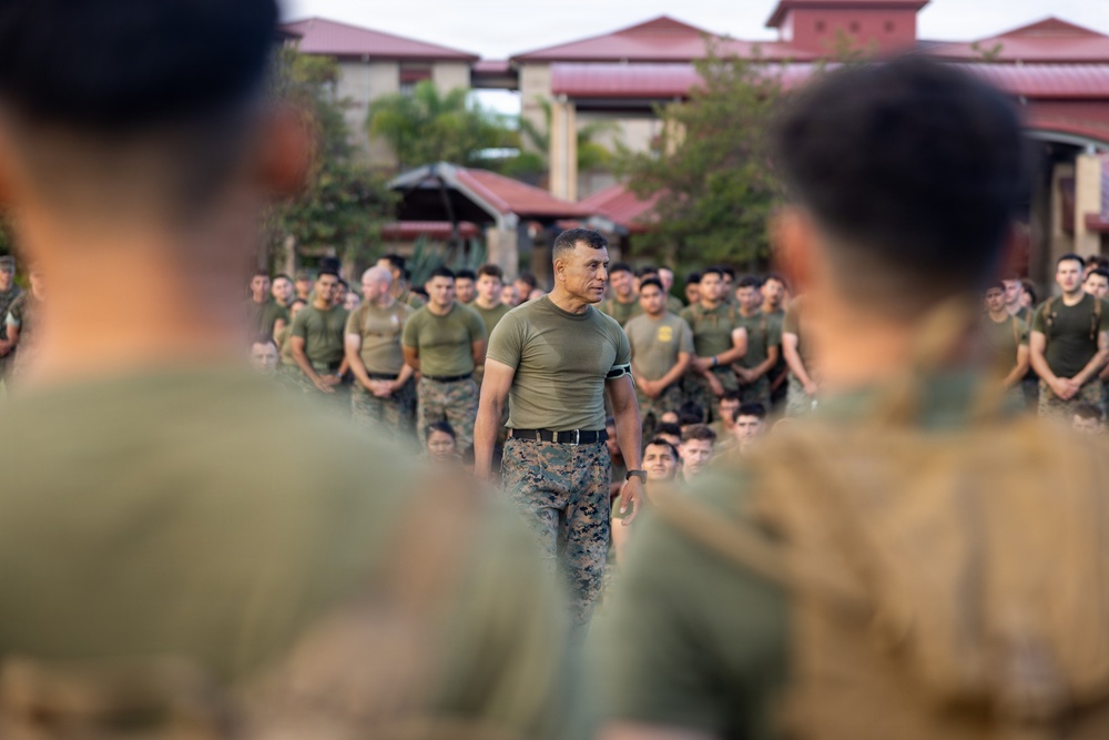 A military formation scene showing a central figure in an olive-green t-shirt and camouflage trousers standing before a large group of uniformed personnel. The central individual appears to be in a leadership or instructional role, addressing the assembled troops. The setting appears to be an outdoor courtyard or parade ground near buildings with red-tiled roofs.