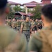 A military formation scene showing a central figure in an olive-green t-shirt and camouflage trousers standing before a large group of uniformed personnel. The central individual appears to be in a leadership or instructional role, addressing the assembled troops. The setting appears to be an outdoor courtyard or parade ground near buildings with red-tiled roofs.