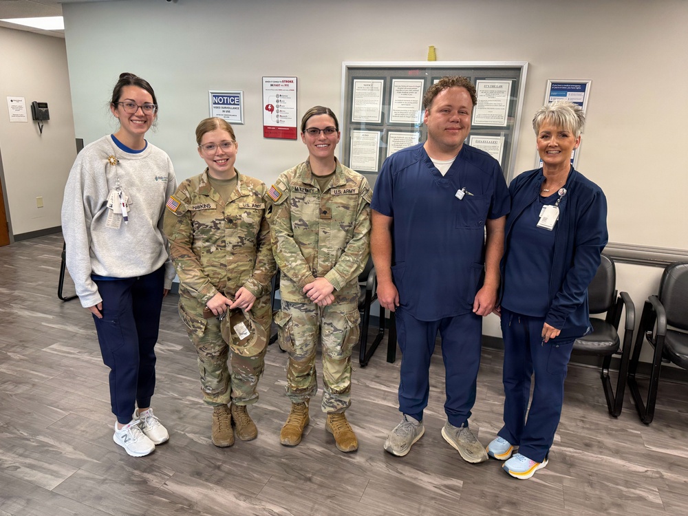 Five individuals are posing together in what appears to be a medical facility waiting area. Two individuals are wearing U.S. Army camouflage uniforms (OCP pattern), while three others are wearing medical scrubs and a sweatshirt with ID badges. The group appears to be gathered for a photo, possibly commemorating a visit or collaboration between military personnel and healthcare staff.