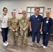 Five individuals are posing together in what appears to be a medical facility waiting area. Two individuals are wearing U.S. Army camouflage uniforms (OCP pattern), while three others are wearing medical scrubs and a sweatshirt with ID badges. The group appears to be gathered for a photo, possibly commemorating a visit or collaboration between military personnel and healthcare staff.
