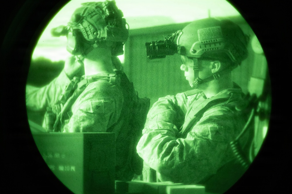 The image is captured through a night vision device, showing two military personnel in full tactical gear including helmets and night vision goggles. The characteristic circular vignette and green monochromatic tones are consistent with Generation 2 or 3 image intensifier night vision optics. The soldiers appear to be in an indoor or enclosed tactical environment.
