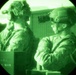 The image is captured through a night vision device, showing two military personnel in full tactical gear including helmets and night vision goggles. The characteristic circular vignette and green monochromatic tones are consistent with Generation 2 or 3 image intensifier night vision optics. The soldiers appear to be in an indoor or enclosed tactical environment.