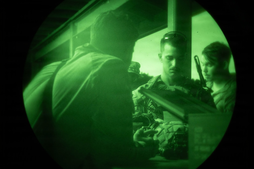 Image captured through a night vision device showing a circular green-tinted field of view. Multiple individuals in military/tactical gear are visible, appearing to be engaged in a tactical operation or training exercise. The characteristic green monochrome rendering and circular vignette confirm this is night vision optic footage.