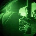 Image captured through a night vision device showing a circular green-tinted field of view. Multiple individuals in military/tactical gear are visible, appearing to be engaged in a tactical operation or training exercise. The characteristic green monochrome rendering and circular vignette confirm this is night vision optic footage.
