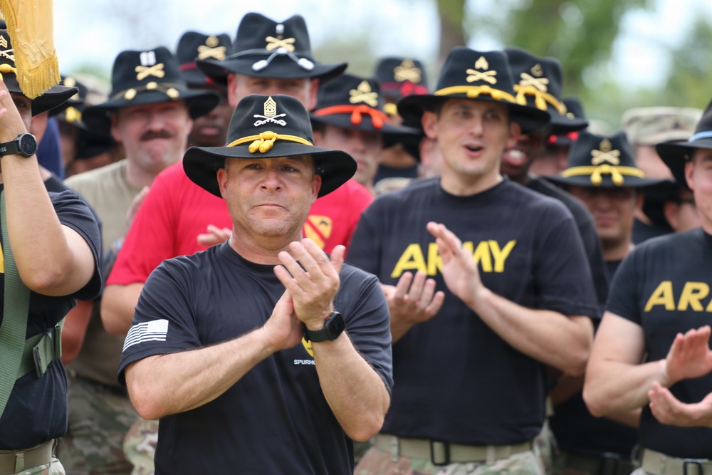 A group of U.S. Army soldiers wearing black cavalry Stetson hats with gold crossed-sabers insignia are gathered at what appears to be a military ceremony or event. The soldiers are dressed in black Army PT (physical training) shirts and are clapping or applauding. The central figure wears a hat with a Command Sergeant Major insignia, indicating senior enlisted rank.