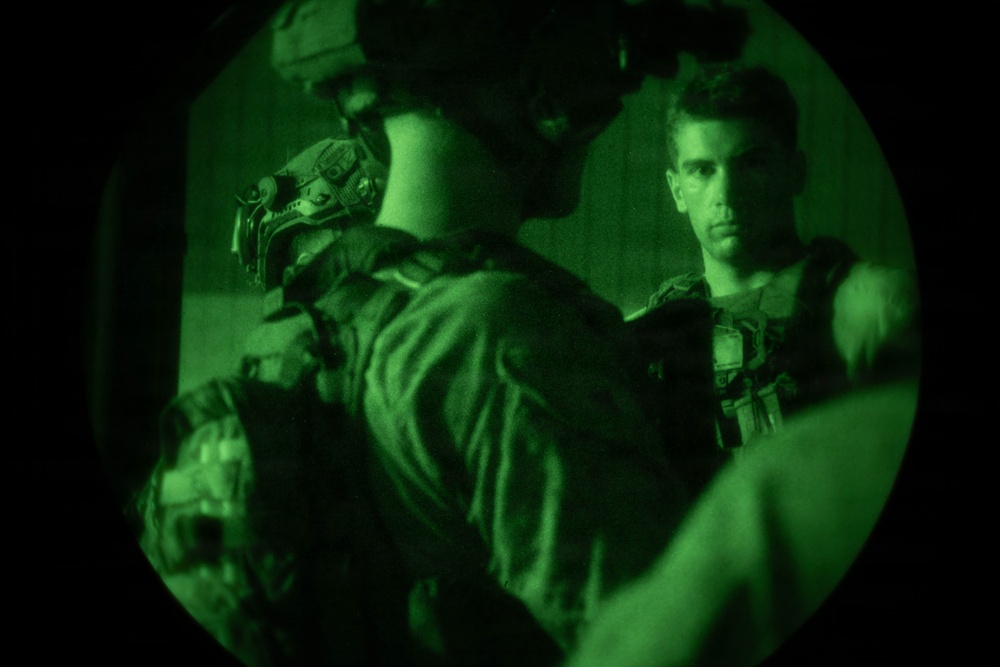 The image is captured through a night vision device, producing a characteristic circular green-tinted view. At least two individuals wearing tactical military gear and body armor are visible in what appears to be an enclosed space. One person faces away from the camera while another faces forward with a direct gaze.