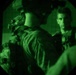 The image is captured through a night vision device, producing a characteristic circular green-tinted view. At least two individuals wearing tactical military gear and body armor are visible in what appears to be an enclosed space. One person faces away from the camera while another faces forward with a direct gaze.