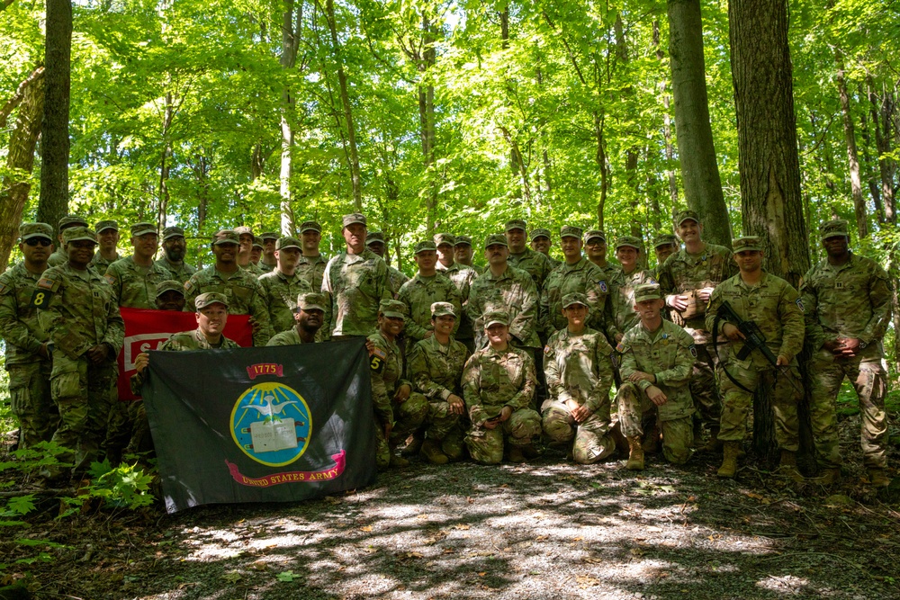 A large group of United States Army soldiers in camouflage uniforms pose for a group photograph in a wooded area. They are holding two flags, including a black United States Army flag dated 1775 and a red unit guidon. The group consists of approximately 25-30 personnel arranged in standing and kneeling rows.