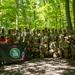 A large group of United States Army soldiers in camouflage uniforms pose for a group photograph in a wooded area. They are holding two flags, including a black United States Army flag dated 1775 and a red unit guidon. The group consists of approximately 25-30 personnel arranged in standing and kneeling rows.