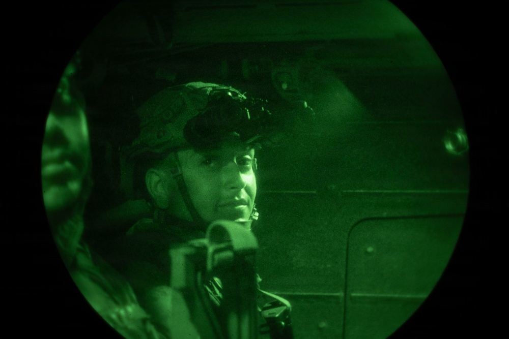 A night vision image captured through a circular lens showing a military personnel wearing a tactical helmet with night vision device (NVD) mounted on it. The image is rendered entirely in green monochrome, characteristic of night vision optics. The subject appears to be inside a military vehicle or aircraft.