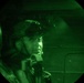 A night vision image captured through a circular lens showing a military personnel wearing a tactical helmet with night vision device (NVD) mounted on it. The image is rendered entirely in green monochrome, characteristic of night vision optics. The subject appears to be inside a military vehicle or aircraft.