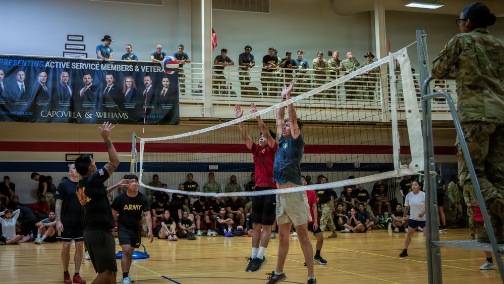 A volleyball game is taking place inside a military gymnasium, with players in Army PT uniforms and civilian attire competing at the net. A large crowd of spectators, including uniformed military personnel, watches from the sidelines and an upper-level balcony. A sponsor banner for 'Capovilla &amp; Williams' law firm, referencing active service members and veterans, is prominently displayed on the upper level.