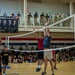 A volleyball game is taking place inside a military gymnasium, with players in Army PT uniforms and civilian attire competing at the net. A large crowd of spectators, including uniformed military personnel, watches from the sidelines and an upper-level balcony. A sponsor banner for 'Capovilla &amp; Williams' law firm, referencing active service members and veterans, is prominently displayed on the upper level.
