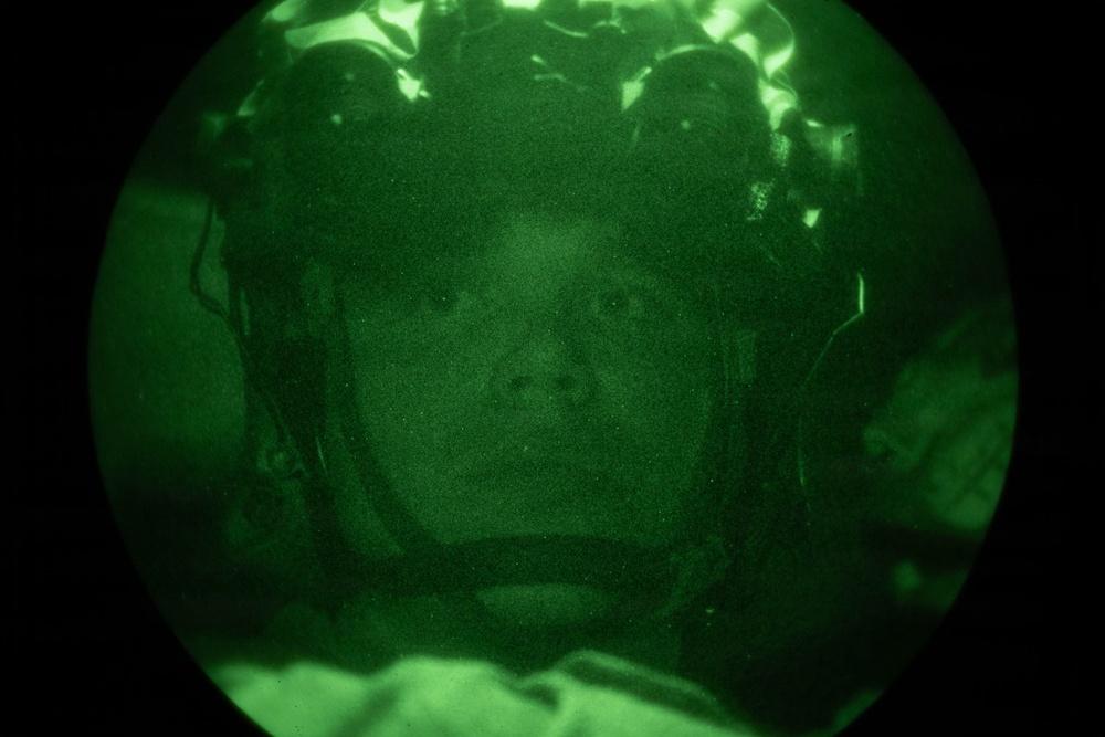 A night-vision image showing a person wearing military or tactical headgear, captured through a night-vision device (NVD) or scope. The circular green-tinted field of view is characteristic of image intensifier tube optics. The subject appears to be wearing a helmet with attached equipment visible at the top of the frame.