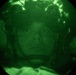 A night-vision image showing a person wearing military or tactical headgear, captured through a night-vision device (NVD) or scope. The circular green-tinted field of view is characteristic of image intensifier tube optics. The subject appears to be wearing a helmet with attached equipment visible at the top of the frame.