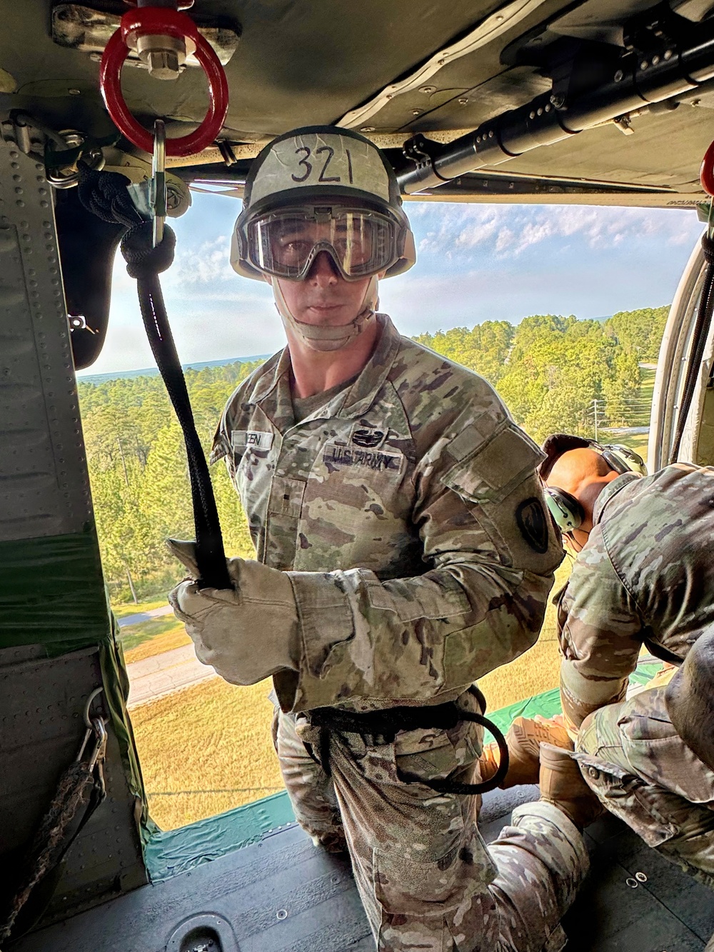 A U.S. Army soldier wearing OCP camouflage uniform, protective goggles, and a helmet marked '321' is positioned inside a military helicopter, holding a rappelling rope connected to a red anchor ring on the ceiling. A second soldier in similar gear is visible to the right, and the open door reveals a forested landscape below, indicating the aircraft is airborne at moderate altitude. The scene depicts a military rappelling or fast-rope training exercise.