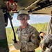 A U.S. Army soldier wearing OCP camouflage uniform, protective goggles, and a helmet marked '321' is positioned inside a military helicopter, holding a rappelling rope connected to a red anchor ring on the ceiling. A second soldier in similar gear is visible to the right, and the open door reveals a forested landscape below, indicating the aircraft is airborne at moderate altitude. The scene depicts a military rappelling or fast-rope training exercise.