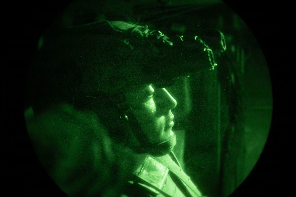 The image is captured through a night vision device, showing a military personnel wearing a tactical helmet with netting/camouflage cover. The circular green-tinted field of view is characteristic of night vision optics. The subject appears to be in profile view, looking to the right, in what appears to be an indoor or enclosed environment.