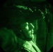 The image is captured through a night vision device, showing a military personnel wearing a tactical helmet with netting/camouflage cover. The circular green-tinted field of view is characteristic of night vision optics. The subject appears to be in profile view, looking to the right, in what appears to be an indoor or enclosed environment.