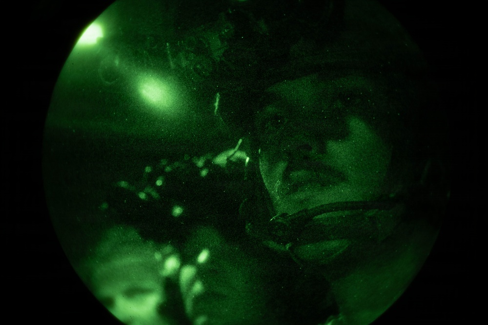 The image is captured through a night vision device, showing a circular green-tinted field of view characteristic of night vision optics. A person wearing military or tactical gear, including a helmet and what appears to be a chin strap or communication equipment, is visible in the frame. The image exhibits the typical grainy, monochromatic green appearance associated with image intensifier night vision technology.