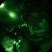 The image is captured through a night vision device, showing a circular green-tinted field of view characteristic of night vision optics. A person wearing military or tactical gear, including a helmet and what appears to be a chin strap or communication equipment, is visible in the frame. The image exhibits the typical grainy, monochromatic green appearance associated with image intensifier night vision technology.