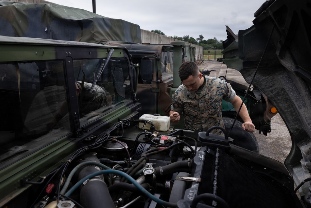 A military service member in camouflage uniform is performing maintenance on the engine of a military tactical vehicle, likely a HMMWV or similar military truck. The scene takes place in what appears to be a motor pool or vehicle maintenance area, with multiple additional military vehicles visible in the background. The open hood reveals a detailed view of the engine compartment.