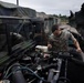 A military service member in camouflage uniform is performing maintenance on the engine of a military tactical vehicle, likely a HMMWV or similar military truck. The scene takes place in what appears to be a motor pool or vehicle maintenance area, with multiple additional military vehicles visible in the background. The open hood reveals a detailed view of the engine compartment.