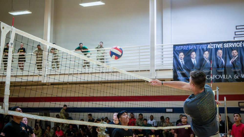 A volleyball game is taking place inside a military gymnasium with a large crowd of spectators, many appearing to be in military uniforms. A player is reaching over or near the net to hit the ball, while teammates and opponents watch closely. A sponsor banner reading 'Representing Active Service' and 'Capovilla &amp; W...' is visible on the upper wall.