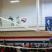 A volleyball game is taking place inside a military gymnasium with a large crowd of spectators, many appearing to be in military uniforms. A player is reaching over or near the net to hit the ball, while teammates and opponents watch closely. A sponsor banner reading 'Representing Active Service' and 'Capovilla &amp; W...' is visible on the upper wall.