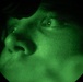 A close-up image of a person's face captured through a night vision device, producing the characteristic green monochromatic appearance. The circular vignette framing is typical of night vision optics. The subject appears to be looking upward with one eye clearly visible.