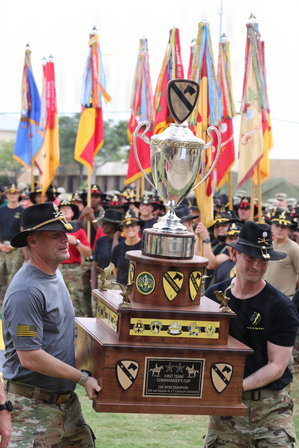 Two soldiers in 1st Cavalry Division attire carry a large ornate trophy called the 'First Team Commander's Cup,' awarded to the CAV Week Champion top battalion in the 1st Cavalry Division. The ceremony takes place in front of a large formation of soldiers wearing cavalry Stetsons and holding unit guidons and colors. The background displays numerous military flags and streamers representing various units.