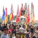 Two soldiers in 1st Cavalry Division attire carry a large ornate trophy called the 'First Team Commander's Cup,' awarded to the CAV Week Champion top battalion in the 1st Cavalry Division. The ceremony takes place in front of a large formation of soldiers wearing cavalry Stetsons and holding unit guidons and colors. The background displays numerous military flags and streamers representing various units.