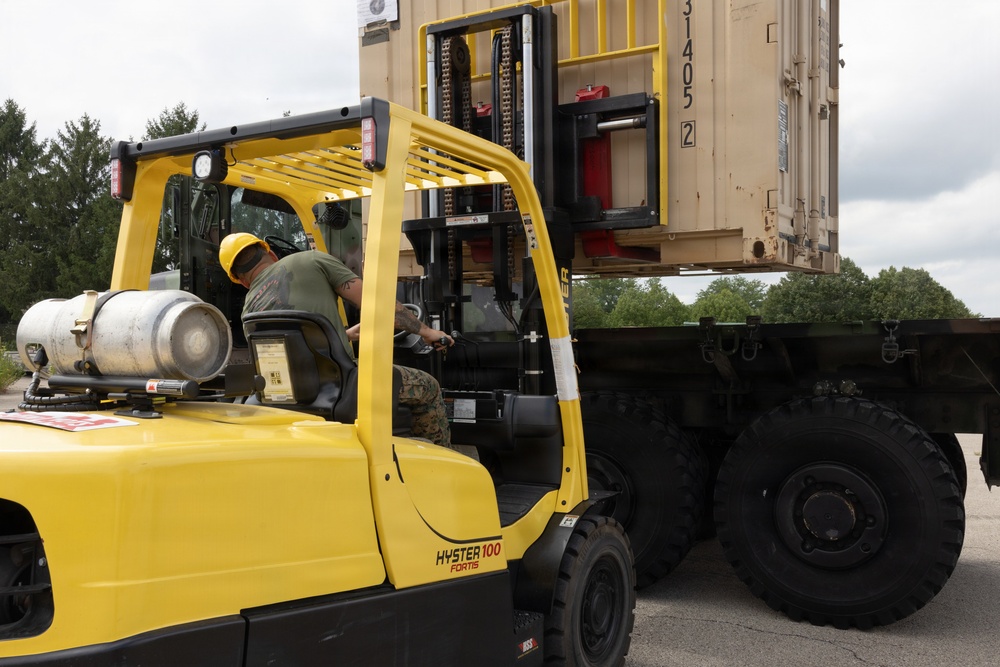 A military personnel wearing a yellow hard hat and camouflage uniform operates a yellow Hyster 100 Fortis forklift. The forklift is actively lifting or positioning a large tan/beige military shipping container onto or off of a flatbed military truck. The operation is taking place outdoors in a paved area surrounded by trees under an overcast sky.