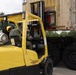 A military personnel wearing a yellow hard hat and camouflage uniform operates a yellow Hyster 100 Fortis forklift. The forklift is actively lifting or positioning a large tan/beige military shipping container onto or off of a flatbed military truck. The operation is taking place outdoors in a paved area surrounded by trees under an overcast sky.
