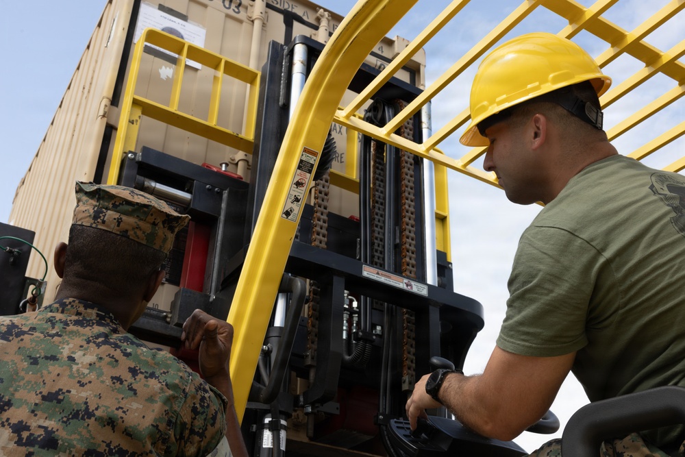Two military personnel are operating a yellow forklift near a large tan shipping container. One individual wearing a yellow hard hat and olive green t-shirt is seated in the forklift operator's position, while another in full camouflage uniform (MARPAT pattern) stands alongside observing or guiding the operation. The scene appears to be a logistics or material handling exercise at a military installation.