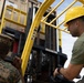 Two military personnel are operating a yellow forklift near a large tan shipping container. One individual wearing a yellow hard hat and olive green t-shirt is seated in the forklift operator's position, while another in full camouflage uniform (MARPAT pattern) stands alongside observing or guiding the operation. The scene appears to be a logistics or material handling exercise at a military installation.