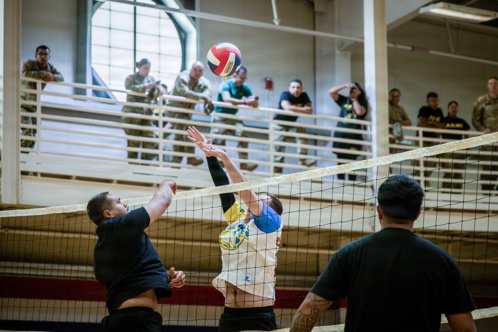 An indoor volleyball game is taking place in what appears to be a military gymnasium or recreational facility. Two players near the net are competing for the ball — one attempting a spike while the other blocks. Spectators wearing military uniforms observe from an elevated balcony in the background.