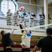 An indoor volleyball game is taking place in what appears to be a military gymnasium or recreational facility. Two players near the net are competing for the ball — one attempting a spike while the other blocks. Spectators wearing military uniforms observe from an elevated balcony in the background.