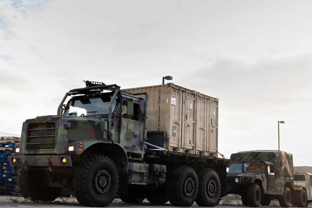 A military convoy scene featuring a large camouflage-painted heavy tactical truck carrying a tan ISO shipping container on a flatbed. A military HMMWV (Humvee) is visible in the background to the right. The scene appears to be at a military installation or staging area during overcast conditions.