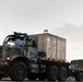 A military convoy scene featuring a large camouflage-painted heavy tactical truck carrying a tan ISO shipping container on a flatbed. A military HMMWV (Humvee) is visible in the background to the right. The scene appears to be at a military installation or staging area during overcast conditions.