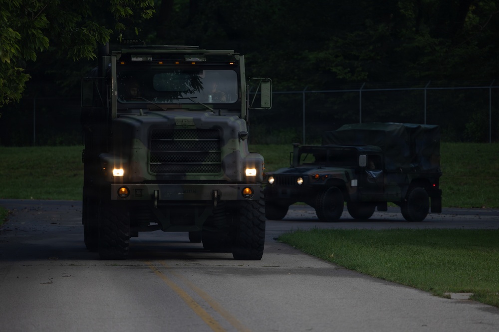 Two military vehicles are traveling in convoy along a paved road during low-light conditions, likely dusk or dawn. The lead vehicle is a large camouflage-painted military truck (consistent with an M939 or similar 5-ton tactical truck) with headlights illuminated, followed by a HMMWV (Humvee). The setting appears to be a military installation or training area, indicated by the chain-link fencing and maintained grounds.
