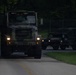 Two military vehicles are traveling in convoy along a paved road during low-light conditions, likely dusk or dawn. The lead vehicle is a large camouflage-painted military truck (consistent with an M939 or similar 5-ton tactical truck) with headlights illuminated, followed by a HMMWV (Humvee). The setting appears to be a military installation or training area, indicated by the chain-link fencing and maintained grounds.